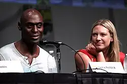Lance Reddick und Anna Torv sitzen am Tisch während einer Panel-Diskussion auf der Comic-Con 2012 in San Diego. Reddick spricht, Torv hört aufmerksam zu. Lance Reddick sitzt auf der linken Seite des Bildes und trägt ein helles Hemd mit einem Kragen. Anna Torv sitzt rechts und trägt ein rotes ärmelloses Oberteil. Beide sitzen hinter einem Tisch, auf dem Mikrofone platziert sind. Vor Anna Torv befindet sich ein Namensschild, auf dem „Lance Reddick“ steht. Der Hintergrund ist unscharf.