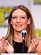 Anna Torv auf der San Diego Comic-Con 2011. Sie steht vor einem Mikrofon und scheint zu sprechen oder eine Präsentation zu halten. Im Hintergrund sind verschwommene gelbe und schwarze Schilder zu sehen, die das Logo der Comic Con darstellen. Anna Torv trägt ein schwarzes Oberteil und hat lange, blonde Haare. Ihre Hände sind erhoben, als ob sie gestikuliert oder etwas erklärt.
