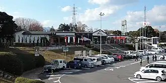 Blick auf einen niedrigen, modernen Gebäudekomplex, der leicht erhöht liegt und von Bäumen umstanden ist. Davor eine Betonfläche mit vielen parkenden Autos, in der Mitte der Fahrzeuge eine freie Fahrgasse mit weißen Pfeilen und einem Rollstuhl-Piktogramm.