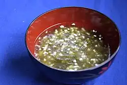 Eine rote Schüssel enthält eine klare Suppe mit Grünalgen und kleinen weißen Würfeln einer weiteren Zutat.