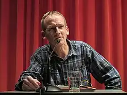 Wolfgang Berends an einem Tisch sithend mit Wasserglas und Mikrofon vor sich