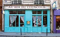 Frontale Farbfotografie eines Restaurants mit hellblauer Fassade, der sich zwischen braunen und violetten Fassaden befindet. Vom Obergeschoss sind nur zwei kleine Balkone zu sehen. Das weiße Reklameschild mit der roten Aufschrift „Au roi du couscous. Specialités marocaines“ geht über die ganze Fassade. Die großen Fenster sind durch Sprossen geteilt und haben weiße Gardinen, in deren Mitte ein Menükasten ist. Oben rechts steht ein Aushängeschild in arabischer Schrift.