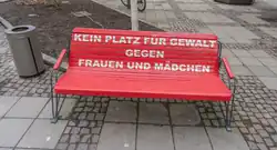 Das Foto zeigt eine Sitzbank, die rot bemalt ist und auf der Lehne in Versalien „KEIN PLATZ FÜR GEWALT GEGEN FRAUEN UND MÄDCHEN“ anzeigt