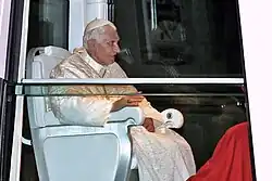 Seitliche Farbfotografie vom Papst in einem geschlossenen Fahrzeug. Er sitzt in einem weißen Stuhl und trägt helle liturgische Kleidung mit Kappe.