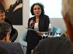 Das Foto ist aus dem Publikum heraus fotografiert, links ist das seitliche Profil einer Person von hinten zu sehen, im Vordergrund und in der rechten Bildhälfte sind ebenfalls sitzende Personen unscharf von hinten abgebildet. Gemma Pörzgen nimmt die Mitte des Bildes ein und sitzt im Fokus auf einem mint-farbenen Stuhl. Sie hält ein Mikrofon in der Hand, trägt einen schwarze Hose, ein weißes Oberteil und einen schwarzen geöffneten Blazer. Ihr Blick geht ins Publikum, zu welchem sie spricht und ihr Haar ist halblang, lockig und dunkel. Im Hintergrund ist der Ausschnitt eines Bildes zu sehen und den größten Teil nimmt eine helle projizierte Fläche ein.