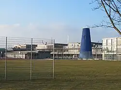 Ein blauer konisch nach oben zulaufender Turm vor niedrigeren Schulgebäuden