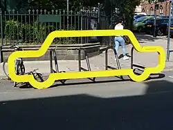 Eine hellgelbe Konstruktion in der Form eines stilisierten Autos, etwa anderthalb Meter hoch und fünf bis sechs Meter breit. Dahinter erkennt man mehrere, parallel aufgestellte Fahrradbügel. Die Konstruktion steht im Rand einer Straße, die Fahrradparkplätze sind durch das gelbe Auto vom Gehweg aus zugänglich. Dahinter sieht man einen kleinen Park und eine städtische Straße.