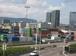 Foto einer Stadt mit einer mehrspurigen Straße im Vordergrund, einem Lagerplatz mit Schiffscontainern dahinter und Hochhäusern im Hintergrund