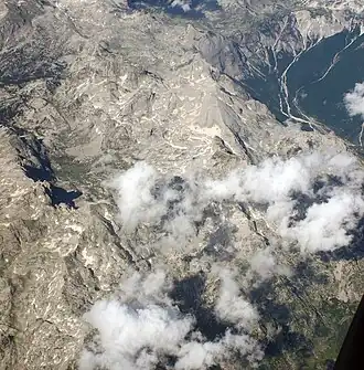 Flugaufnahme von Südwesten mit der Spitze rechts der Mitte über dem Wolkenband zwischen dem Shala-Tal rechts und dem Polje Livadhet e Bogës links