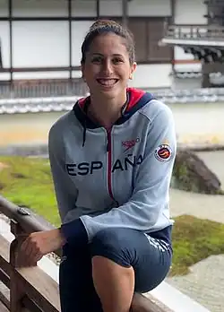 Toledano sitzt auf einem Balkongeländer. Sie trägt eine blaue Trainingshose und eine graue Jacke mit der Aufschrift ESPANA.