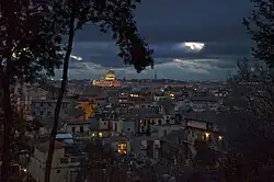 Blick auf die Stadt bei Nacht mit einem angestrahlten Dom. In einzelnen Häusern ist Licht zu sehen und ein heller Mondschein dringt durch die Wolken hervor. Im Vordergrund sind die Silhouetten von Bäumen zu erkennen.