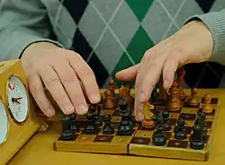 Auf einem hölzernen Tisch liegt aufgeklappt ein sehr kleines Steckschachbrett von der Größe eines Taschenbuches, es ist wie die Figuren aus hellem und sehr dunklem Holz gefertigt. Eine Erhöhung der schwarzen Felder ist nicht zu erkennen, die schwarzen Figuren sind oben mit Nagelköpfen markiert. Am linken Bildrand, direkt neben dem Schachbrett, steht eine hölzerne Schachuhr mit runden Zifferblättern. Hinter dem Schachbrett sitzt uns gegenüber ein Schachspieler. Er sitzt ganz nah am Tisch und man sieht nur wenig von seinem graublauen Pullover mit Rautenmuster. Das Bild zeigt in Großaufnahme, wie er mit den Fingern beider Hände die auf dem Brett verteilten Figuren betastet.