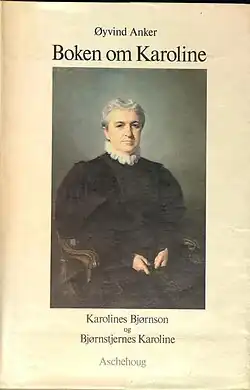 Das abgelichtete Buchcover zeigt den Titel „Boken om Karoline“, geschrieben von Øyvind Anker. Außerdem ist ein Gemälde einer älteren Frau auf einem Stuhl abgebildet. Sie trägt dunkle Kleidung und hat weiße Haare. Ihre Hände ruhen im Schoß, was eine ruhige und gelassene Haltung vermittelt. Unterhalb des Gemäldes steht der Text „Karolines Bjørnson og Bjørnstjernes Karoline“, der den Bezug zu Karoline und Bjørnstjerne Bjørnson hervorhebt. Ganz unten ist das Wort „Aschehoug“ sichtbar, was vermutlich der Name des Verlags ist.