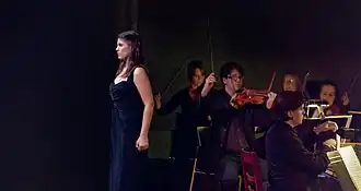 Frau mit langen dunklen Haaren in dunkelblauem Kleid steht nach rechts gewandt vor schwarzem Hintergrund, hinter ihr halten Musiker mit Streichinstrumenten ihre Streichbögen hoch, ganz links sitzt ein Pianist.