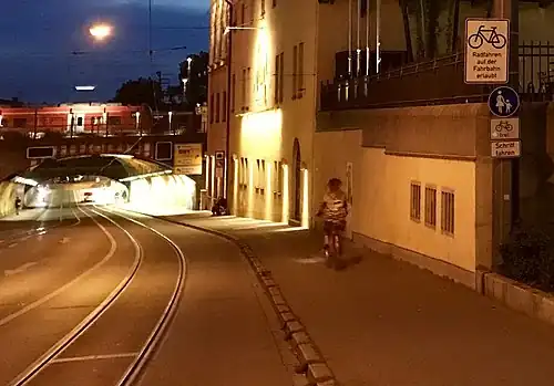 Eine lange Bahnunterführung in Augsburg mit leichtem Gefälle, glänzende Meterspur-Straßenbahnschienen mit etwa 1,50&nbsp;m Abstand vom Bordstein, Überholverbot (durchgezogene weiße Linie) für Autos in der beleuchteten Unterführung nahe dem Hauptbahnhof mit einem rot-weißen Zug oberhalb der leicht gebogenen Tunnelüberwölbung. Eine Fahrradfahrerin (vermutlich) auf dem etwa 2,50 Meter breiten Gehweg rechts, ein Bettler sitzt ein wenig weiter unten am Rand an der hohen Hauswand. Verkehrsschild Fussgänger mit Zusatzschild Fahrradfahren frei und Hinweistafel „Radfahren auf der Fahrbahn erlaubt“. Links auf der gegenüberliegenden Straßenseite trennt keine Wand mehr den Bürgersteig von der aufwärtsführenden Fahrbahn ab. Blaue Stunde am Abend mit Gebäude einer Brauerei rechts im Bild.