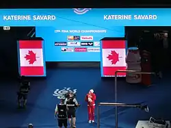 Farbfoto von Katerine Savard im roten Trainingsanzug mit weißer Bademütze. Sie ist nur klein im Vordergrund zu sehen, im Hintergrund eine riesige Medienwand mit den Namen diverser Sponsoren eingerahmt von der kanadischen Flagge.