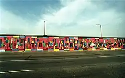 Frontale Farbfotografie von der Berliner Mauer mit einem Bild, das von der gegenüberliegenden Straßenseite aufgenommen wurde. Abstrakte Figuren sind in kleine, bunte Rechtecke gemalt, die auf rotem Untergrund sind. Über und unter dem Bild reihen sich bunte Querbalken aneinander.