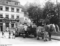 Das Bild zeigt eine mit Paketen schwer beladene stehende Kutsche, die von zwei Pferden gezogen wird. Hinter ihr befindet sich ein Haus mit dem Schriftzug „Zur letzten Instanz“