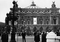 Das Bild zeigt die Vorderfassade der Opéra Garnier in Paris, einer bekannten Oper in Frankreich. Die Architektur des Gebäudes ist aufwändig gestaltet, mit reich verzierten Statuen, Säulen und detaillierten Ornamenten. Besonders auffällig auf diesem Bild ist, dass Nazi-Flaggen das Gebäude schmücken, was darauf hinweist, dass das Foto während der deutschen Besatzung von Paris im Zweiten Weltkrieg aufgenommen wurde. Im Vordergrund sind Menschen zu sehen, einige in Militäruniformen, andere in ziviler Kleidung. Die Szene vermittelt eine Mischung aus kultureller Pracht und der düsteren Realität der Besatzung.