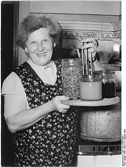 lächelnde ältere Frau mit Einkochgläsern auf dem Topfeinsatz; im Hintergrund der zugehörige verzinkte große Einkochtopf (1954)