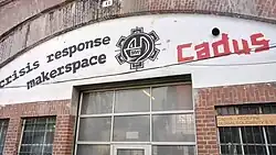 Eingangsbereich eines Backsteingebäudes mit großem Schriftzug „Cadus Crisis Response Maker Space“ und Logo über den Flügeltüren