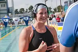 Leverenz im schwarzen Badeanzug und mit silberner Bademütze steht unter freiem Himmel neben dem Pool. Sie trägt ein schwarzes Headset und wird gerade interviewt.