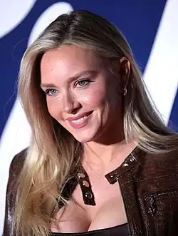 Camille Kostek mit langen, blonden Haaren und blauen Augen. Sie trägt eine braune Lederjacke trägt. Die Jacke hat Reißverschluss- und Knopfdetails. Camille Kostek lächelt zum Fotografen. Im Hintergrund ist ein blauer Farbton mit einem weißen Muster zu sehen.