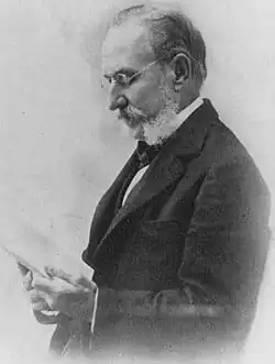 Lesender Mann im gehobenen Alter im Seitenprofil, nach links schauend ab Becken im dunklen Anzug, weißem Hemd und dunkler Krawatte mit einem Blatt Papier in der Hand in schwarzweiß.