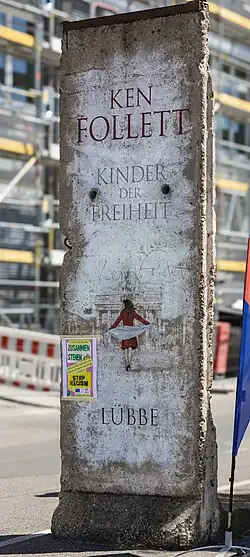 Ein Mauersegment der Berliner Mauer, Beschriftung Ken Follett Kinder der Freiheit, Lübbe. Mauer ist leicht weiß getüncht, Illustration einer Frau in rotem Kleid vor dem Brandenburger Tor