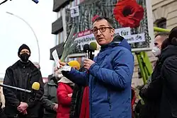 Cem Özdemir spricht mit zwei Mikrofonen in der linken Hand. In der rechten Hand hält er einen Porree und einen Notizzettel. Im Hintergrund sind einige Personen und ein Plakat an einem Traktor zu sehen.