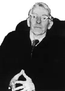 In einem Stuhl mit schwarzer Rückenlehne sitzender älterer Mann in einem schwarzen Anzug oder einer schwarzen Robe, weißem Hemd mit Krawatte ab Becken aufwärts in schwarzweiß von vorne in die Kamera schauend.