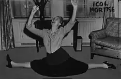 Charlotte Greenwood, die einen Spagat am Boden ausführt. Sie trägt eine Jacke und einen Rock, kombiniert mit Absatzschuhen. Die Arme sind in die Luft gestreckt, wobei in einer Hand ein Taschentuch gehalten wird. Im Hintergrund sieht man ein Zimmer mit Möbeln. Es gibt einen Tisch, auf dem eine Vase mit Blumen steht, einen Sessel und einen Heizkörper unter einem Fenster. An der rechten Wand des Bildes ist handschriftlich „ICOI MORTISE“ geschrieben.