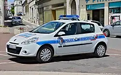 Foto eines Einsatzfahrzeugs der Police municipale Châteauneuf-sur-Charente, aufgenommen von schräg vorne, so dass Vorder- und Fahrerseite sichtbar sind. Der viertürige Pkw vom Typ Renault Clio III ist weiß lackiert und hat eine blaue Bauchbinde. Diese ist an der Fahrzeugseite im Bereich der Türen mit dem Schriftzug "Police municipale" in Versalien und blauen Lettern versehen. Auf der Fahrertür ist zudem das frankreichweit einheitliche Wappen der Police municipale angebracht, das dem Armabzeichen auf der Uniform entspricht. Auch auf der Fahrzeugvorderseite ist der Schriftzug "Police nationale" angebracht, in blau auf weißem Grund und eingefasst von einem roten Rechteck. Auf dem Dach des Autos ist ein Warnbalken mit Blaulicht installiert. Das Fahrzeug ist geparkt, offenbar auf einem Platz. Im Hintergrund befindet sich eine Straße mit Ladengeschäften.