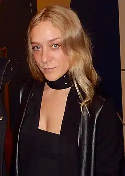 Chloë Sevigny mit blonden, gewellten Haaren, hat blondes Haar und trägt ein schwarzes Outfit, das einen schwarzen Pullover mit tiefem Ausschnitt und eine schwarze Jacke umfasst. Die Jacke hat auffällige, lederähnliche Elemente an den Schultern, die ihr einen modernen und stilvollen Look verleihen. Im Hintergrund sind keine weiteren klaren Details oder Elemente zu erkennen.