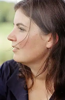 Auf dem Porträt blickt Christa Pfafferott nach links, sie ist im Profil zu sehen. Sie trägt eine dunkelblaue Bluse. Eine dunkelbraune Strähne ihres offenen, mittellangen Haares wird ihr vom Wind ins Gesicht geblasen.