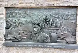 Metallrelief mit leichten, dreidimensionalen Elementen, Tom Pryce im Porträt mittig dargestellt, links und rechts zwei Shadow-Formel-1-Fahrzeuge, im Hintergrund eine Rennstrecke und eine Hügellandschaft mit Wäldern