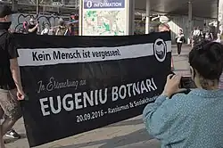 Zwei Personen halten ein schwarzes Transparent auf einem größeren Platz. Dahinter stehen und bewegen sich weitere Personen, davor fotografiert eine Person das Transparent auf dem steht „Kein Mensch ist vergessen! In Erinnerung an Eugeniu Botnari. 20.09.2016 – Rassismus & Sozialchauvinismus tötet!“