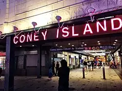 Dämmerungsbild. Oben beleuchtete helle Fassade mit Ornamenten, mittig große beleuchtete Lettern CONEY ISLAND, unten Menschen in und vor einer beleuchteten Fußgängerpassage, die als Ladenzeile und Zugang zum Bahnhof dient.