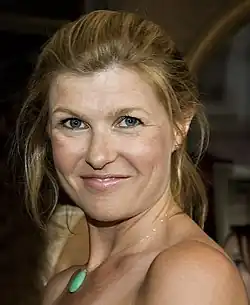 Connie Britton mit blonden, lockeren Wellen im Haar und lächelt. Sie trägt ein schlichtes, elegantes Outfit und eine auffällige Kette mit einem grünen Anhänger, der im Mittelpunkt des Bildes steht. Im Hintergrund ist eine Innenraumszene zu erkennen, einschließlich eines Spiegels und einiger unscharfer Objekte. Der Fokus liegt auf der Frisur und dem Schmuck, wobei die Details der Kette und die sanften Wellen der Haare besonders hervorgehoben werden. Die Lichtverhältnisse lassen die Szene warm und einladend wirken.