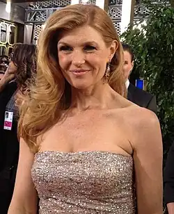 Connie Britton in einem schulterfreies, funkelndes Abendkleid, lächelt in die Kamera. Das Kleid scheint aus einem glitzernden Material wie Pailletten gefertigt zu sein und passt sich eng an die Körperform an, wodurch die Silhouette betont wird. Es reflektiert das Licht, wodurch ein glamouröser Effekt entsteht.Conni Britton hat langes, welliges Haar, das auf eine Seite gestylt ist, was zu ihrem eleganten Aussehen beiträgt. Im Hintergrund des Bildes sind weitere Personen zu erkennen, die teilweise sichtbar und ebenfalls formell gekleidet sind. Außerdem befindet sich eine grüne Pflanze im Hintergrund, die etwas Natürlichkeit in die Szene bringt.