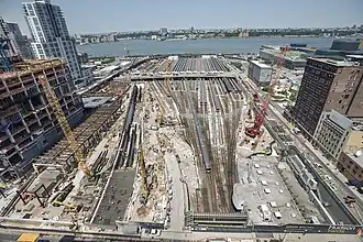Schrägaufnahme einer Gleisharfe, rechts und links umgeben von unterschiedlich hohen Gebäuden, hinten vom Hudson River. Straßenbrücke queren rechts nach links die Gleise, es sind Baukräne und Rohbauten aus Stahl zu sehen. Diesiges Sonnenlicht, alles wirkt industriell und die Farben ausgewaschen mit viel Grau.