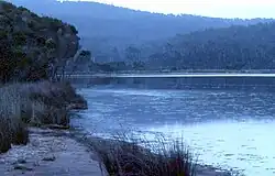 Das Foto eines Sees. Der Betrachter steht am Ufer des Sees an einer sandigen Stelle, auf der einige Gräser wachsen. rechts von Ihm liegt der See, der in etwa zwei Drittel der Fläche des Bildes bis zum Horizont einnimmt. Am anderen Ufer des Sees befinden sich bewaldete Hügel.