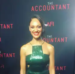 Cynthia Addai-Robinson in einem ärmelosen, glitzernden grünen Kleid mit offenen Rücken lächelt bei der Premiere des Films "The Accountant" 2016. Sie steht vor einem Hintergrund steht, auf dem mehrfach der Schriftzug „THE ACCOUNTANT“ in großen roten Buchstaben zu sehen ist. Zusätzlich ist der Text „AFI“ in roten Buchstaben auf dem Hintergrund abgebildet.