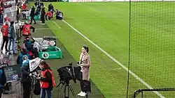 Das Foto wurde aus dem obigen Zuschauerraum auf das Spielfeld fotografiert und zeigt die abwartende Esther Sedlaczek auf dem Seitenstreifen des Fußballfeldes, der Mercedes-Benz-Arena beim DFB-Pokal-Spiel VfB Stuttgart gegen BVB Dortmund, vor einer Stativkamera ein Mikrofon mit beiden Händen vor dem Körper haltend. Sie trägt einen langen beigen Trenchcoat und weiße Turnschuhe. Haare zu einem Pferdeschwanz zusammengebunden. Weiter ist reges Treiben im Hintergrund von Kameramännern und Ordner und Pressemitarbeitern neben dem Spielfeldrand zu sehen.