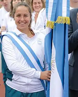 Rocío Sánchez Moccia trägt eine weiße Trainingsjacke mit einer blau-weiß-blauen Schärpe, mit der rechten Hand hält sie die argentinische Flagge.