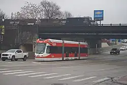 Straßenszene unter grauem Himmel. Straßenbahn, Kraftfahrzeuge, darüber Eisenbahnbrücke mit Güterzug.