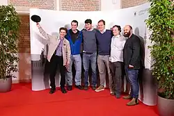 Sechs Männer in legerer Kleidung stehen eng nebeneinander vor einem Diorama der Stadt München.