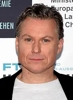 Dirk Borchardt, mit kurzem, gewelltem Haar und neutralem Gesichtsausdruck, trägt eine schwarze Jacke über einem weißen Hemd. Der Hintergrund enthält Text in deutscher Sprache, wie "DEUTSCHE AKADEMIE" und andere teilweise sichtbare Wörter.