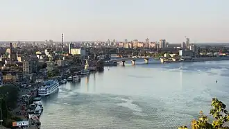 Von der rechten unteren Fotoecke bis zur Mitte erstreckt sich der Fluss. Daran schmiegt sich die Stadt halbkreisförmig von unten links nach rechts. Einige Schiffe und Fabriken sind links zu erkennen, in der Mitte befindet sich eine Autobrücke, und in der Ferne sind Wolkenkratzer.