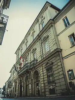 Das Teatro Fraschini in Pavia, Italien, ist ein beeindruckendes Beispiel neoklassizistischer Architektur. Die Fassade des Gebäudes besteht aus Stein und ist reich verziert. Über dem Haupteingang befinden sich drei Bögen, die von eleganten Säulen getragen werden. Ein Balkon mit einem kunstvoll geschmiedeten Geländer erstreckt sich über die Bögen. Die Fassade ist mit drei Flaggen geschmückt: der italienischen Nationalflagge, der Flagge der Stadt Pavia und einer weiteren Flagge. Die Fenster des Gebäudes sind groß und mit dekorativen Rahmen versehen. Das Theater hat mehrere Stockwerke, die durch ihre symmetrische Gestaltung auffallen. Die gesamte Struktur wirkt gut erhalten und gepflegt. Vor dem Gebäude gibt es eine Straße, auf der einige Menschen spazieren gehen.